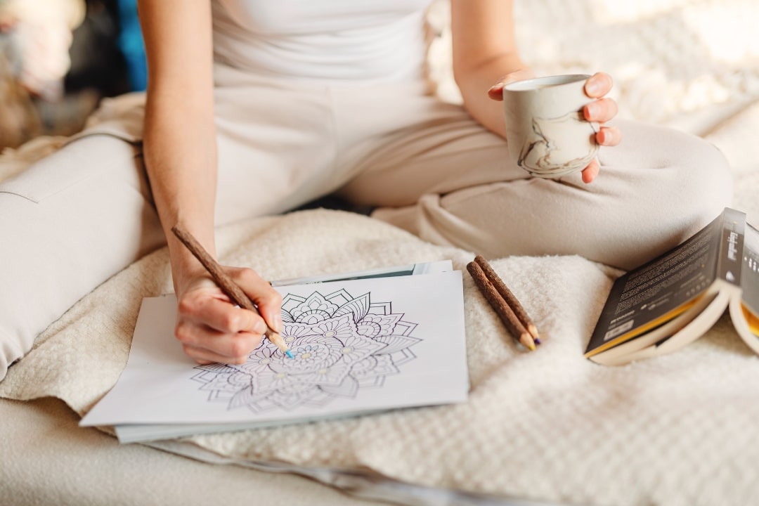 Person sitzt im Schneidersitz mit einer Tasse Tee auf einer weichen Decke und malt Mandala-Bilder aus