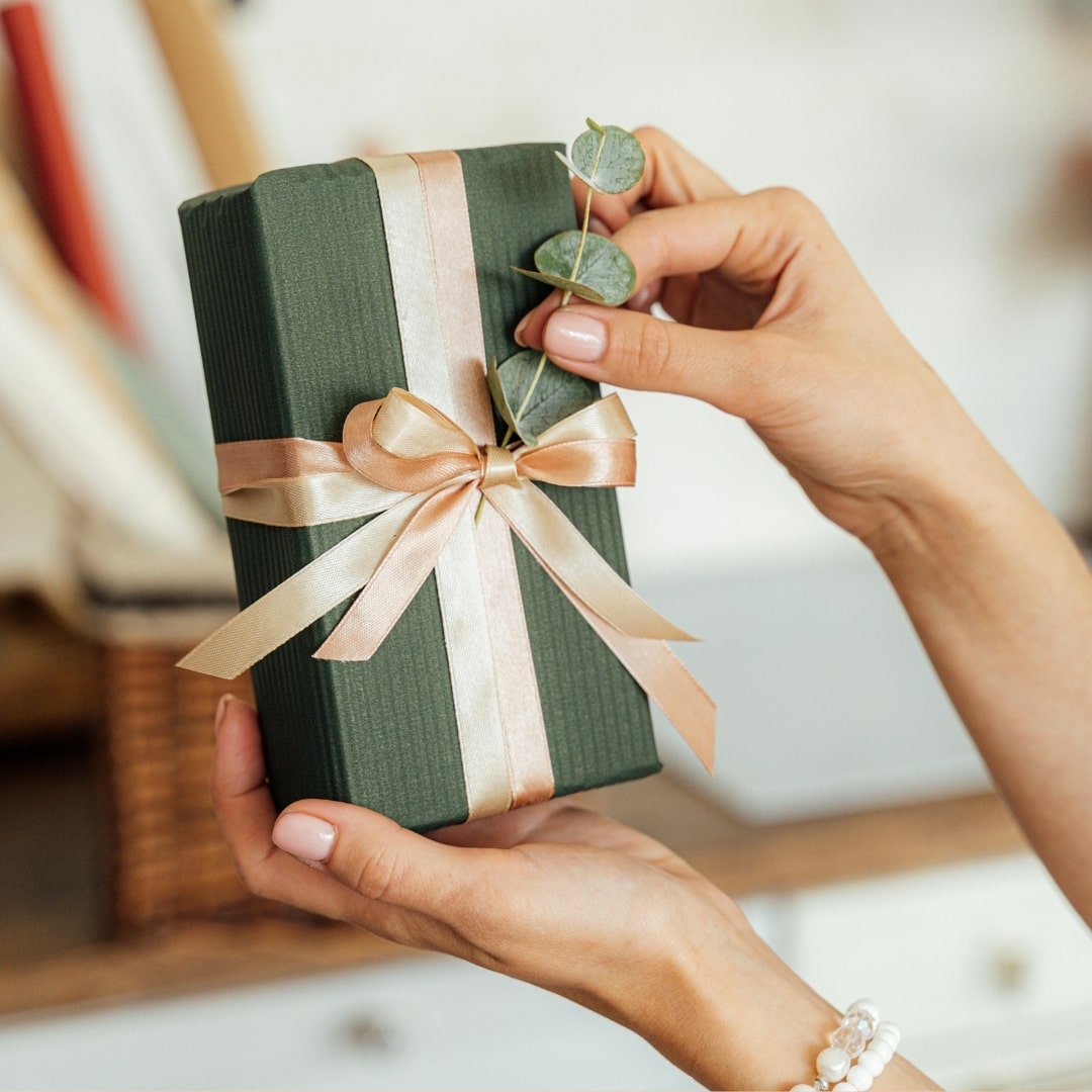 Hände mit verpacktem Geschenk mit Schleife und Eukalyptuszweig als Dekoration.