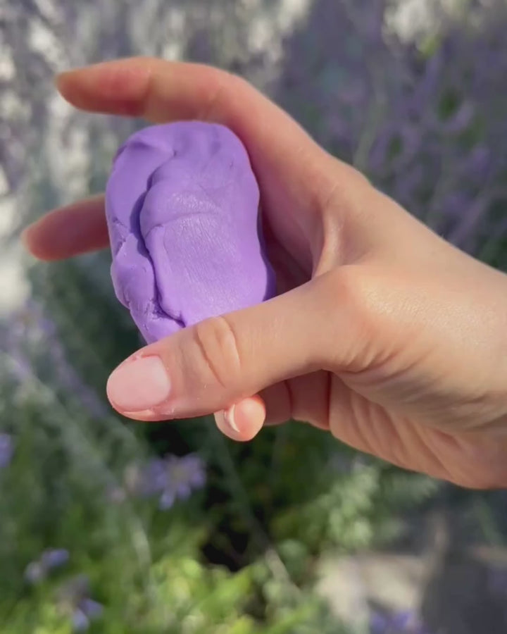 Eine Hand, die die lilafarbene Knete "Zendou Relax" sanft knetet, drückt und formt. Die Bewegungen sind langsam und veranschaulichen die weiche Textur der Knete. Im Hintergrund sind Lavendelpflanzen zu sehen, die die beruhigende Atmosphäre sowie die Nähe zur Natur unterstreichen.