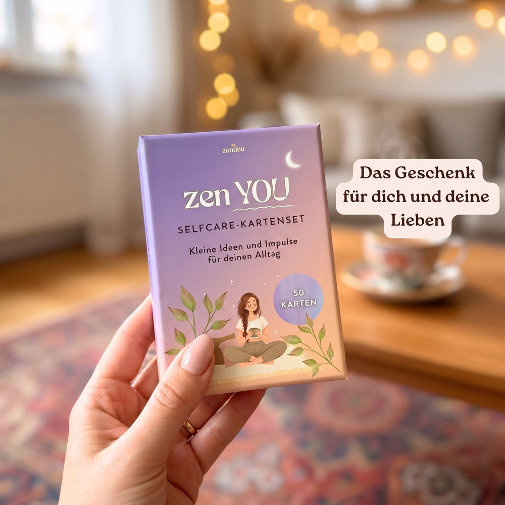 Hand hält das Zen YOU Selfcare-Kartenset in einem gemütlichen Wohnzimmer mit Lichterkette und Teetasse