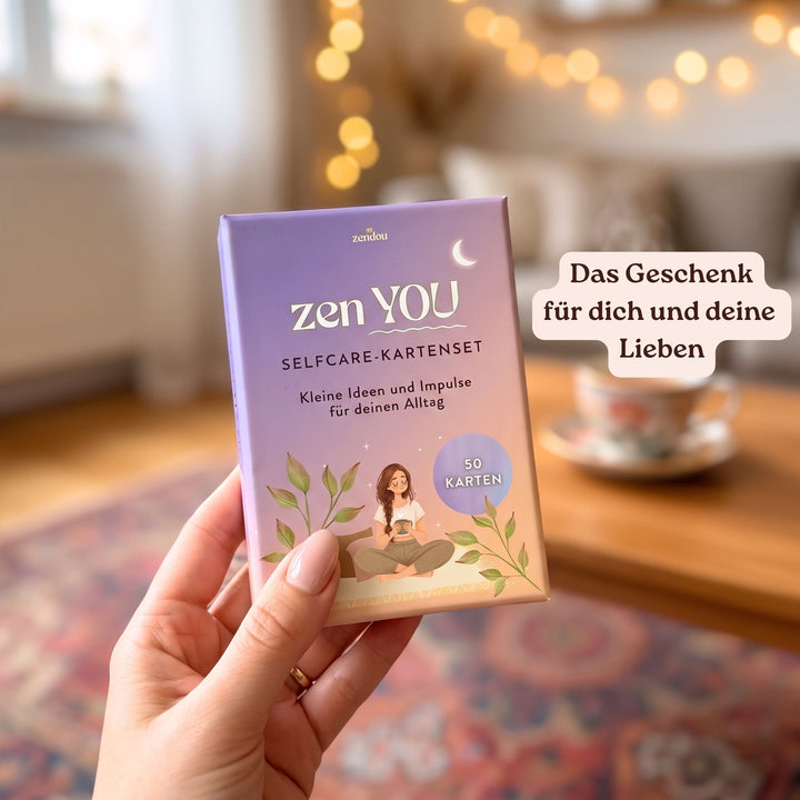 Hand hält das Zen YOU Selfcare-Kartenset in einem gemütlichen Wohnzimmer mit Lichterkette und Teetasse